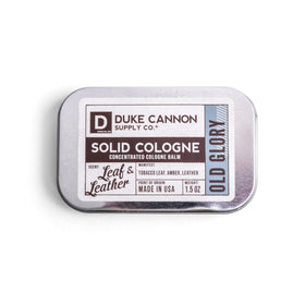 Duke Cannon Old Glory solid cologne tin label showcases premium tobacco-amber-leather scent for quick refresh.