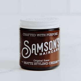 Hands dispensing SAMSON'S HAIRCARE Matte Styling Cream for even coverage.